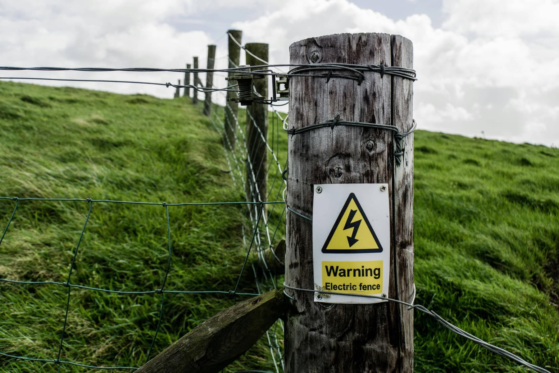 A wooden fence post with a yellow electric fence warning sign in a green field